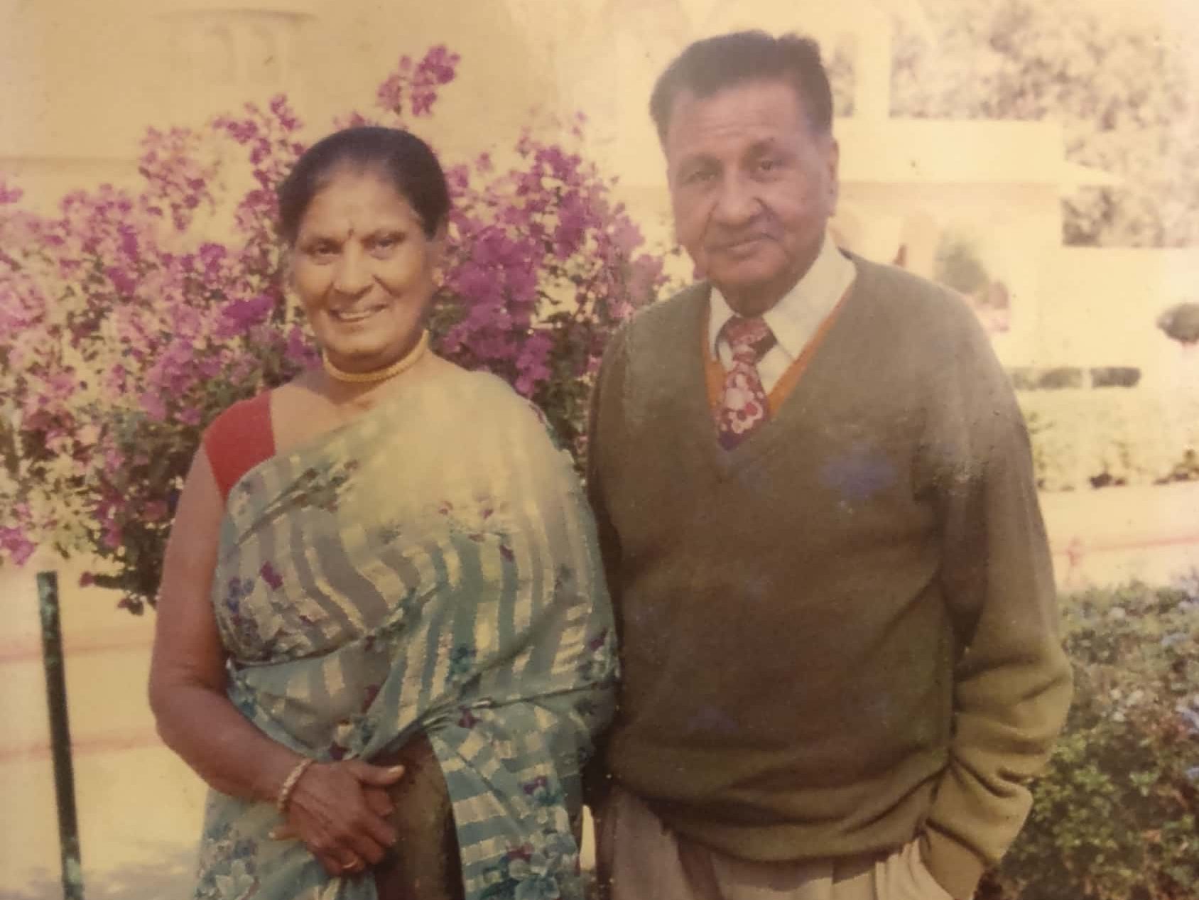 An elderly couple standing together outdoors, smiling. The woman is wearing a traditional saree with a floral design, while the man is dressed in a sweater and tie. Bright purple flowers in the background add color to the scene.