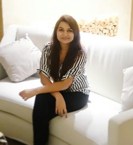 A young woman sitting on a white couch, wearing a black and white striped shirt and black pants, smiling at the camera with pillows in the background.