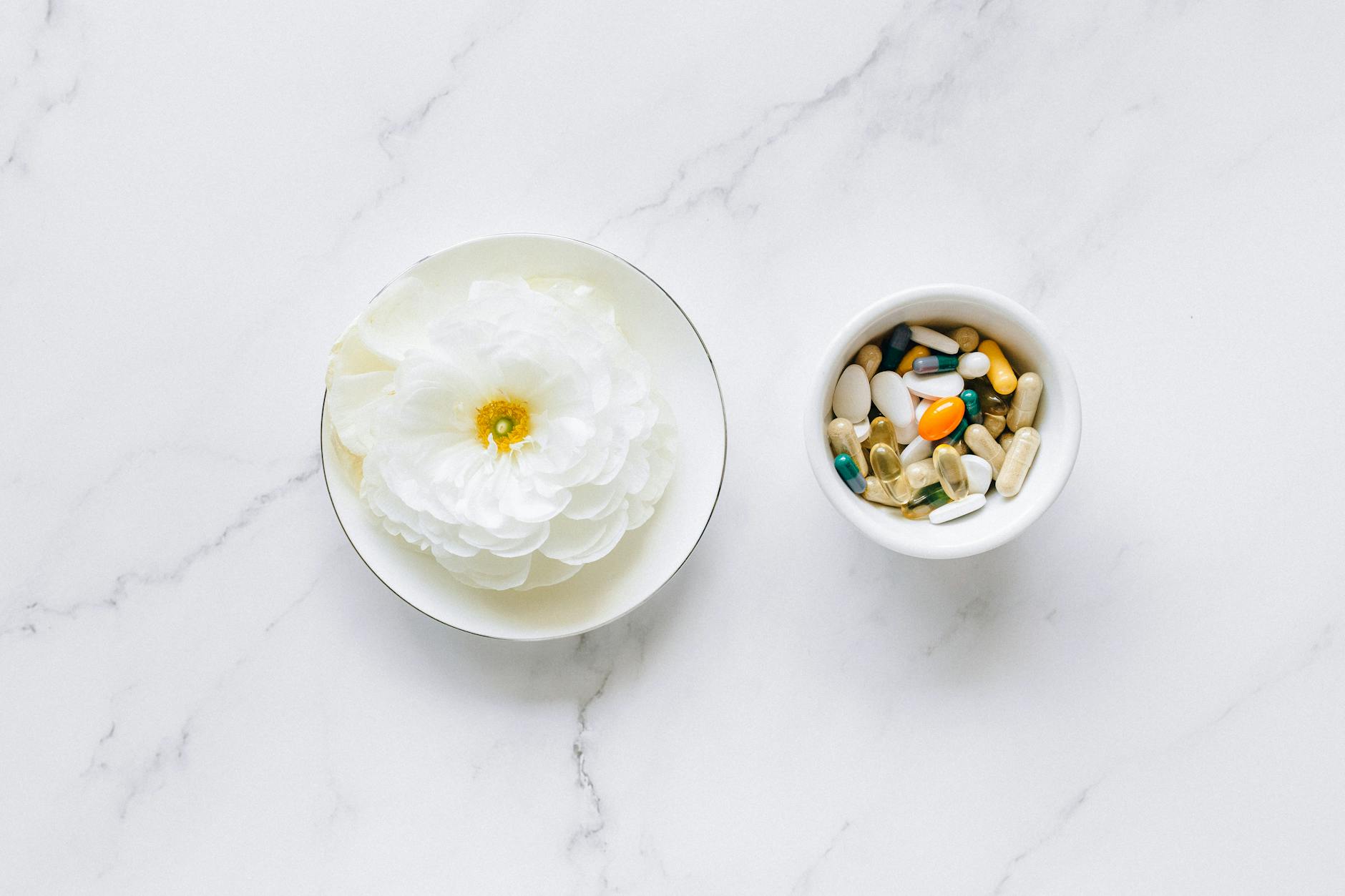 A white flower in a small dish next to a bowl filled with various vitamins and supplements on a marble surface.
