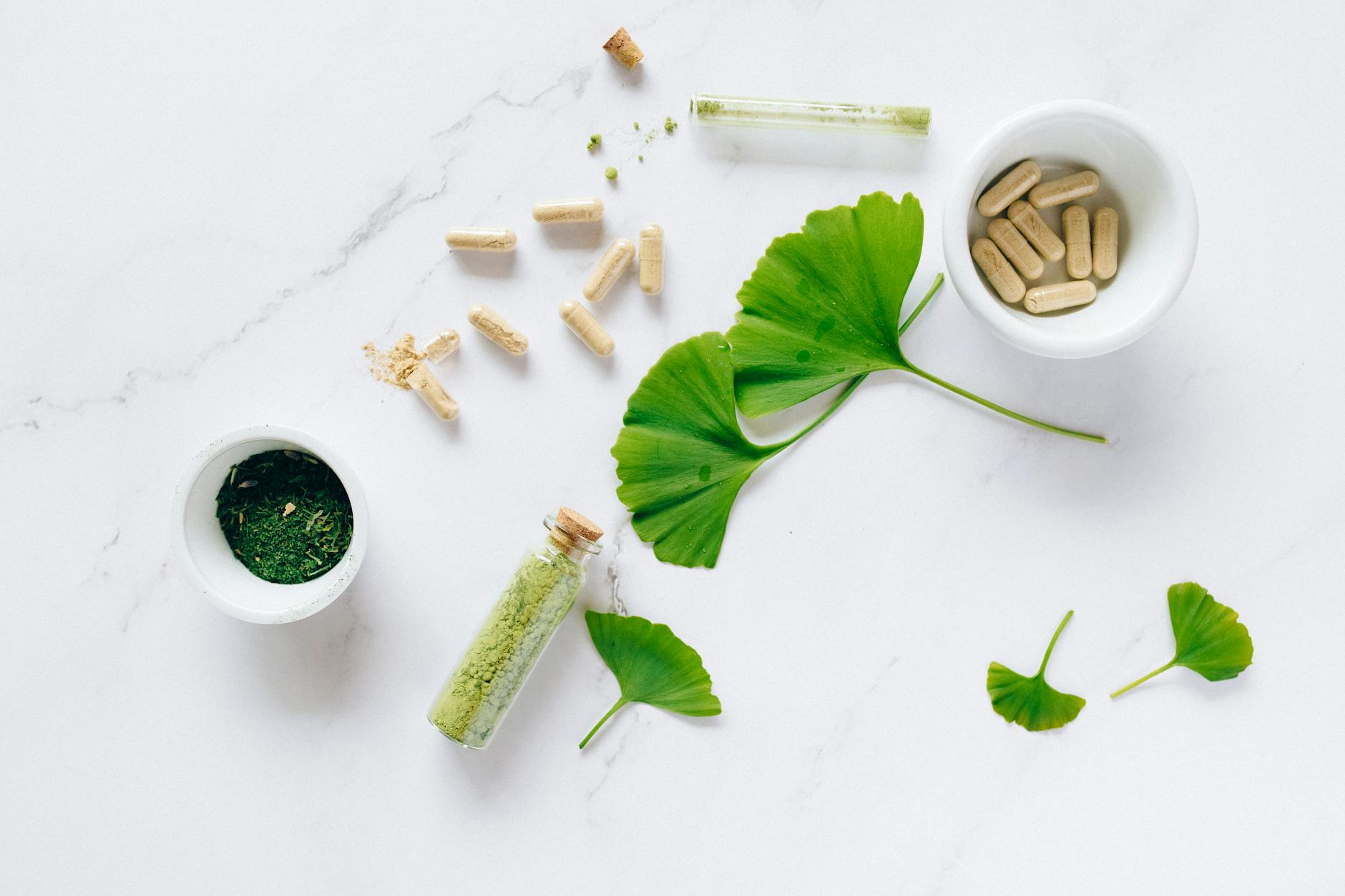 An arrangement of herbal supplements on a marble surface, including green powder in a small vial, a bowl of capsules, and green leaves.